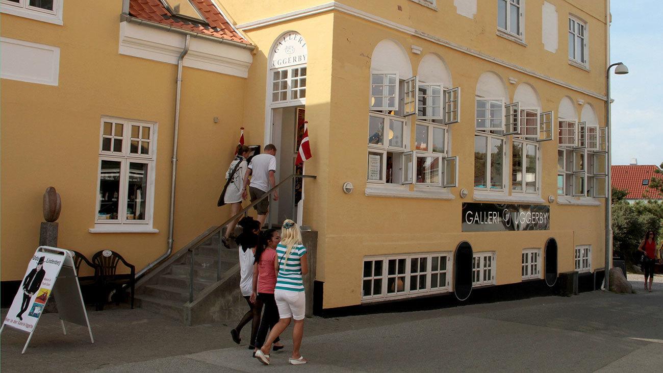 Besøgende går ind i en galleribygning med gule vægge og store vinduer i en by.