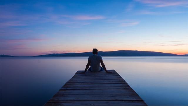 En person sidder på en træbro ved vandet ved solnedgang, omgivet af stille landskab og bløde farver.
