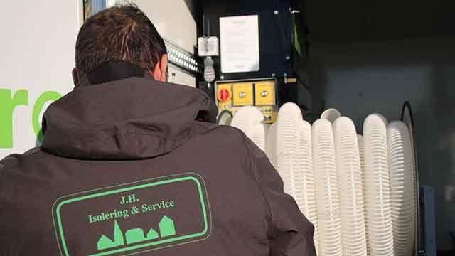 En mand i en jakke med logoet "J.H. Isolering & Service" står foran en maskine med slanger.