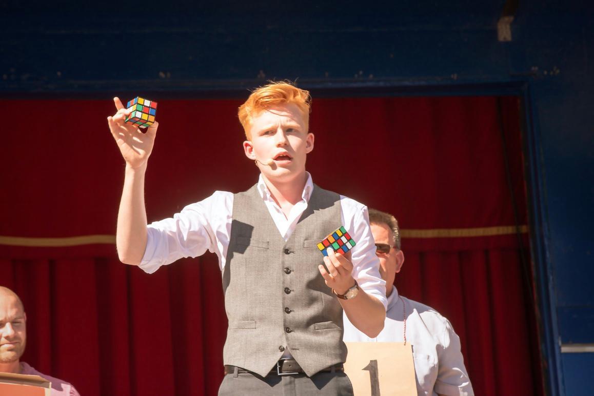 En ung mand med rødt hår holder to Rubiks terninger op, mens han står på en scene med en rød baggrund.