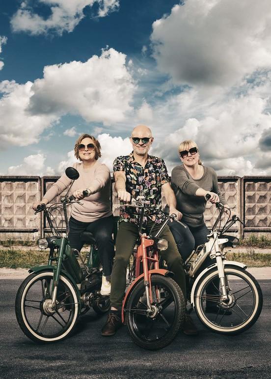 Tre personer står på elcykler med farverige rammer under en dramatisk skyet himmel.