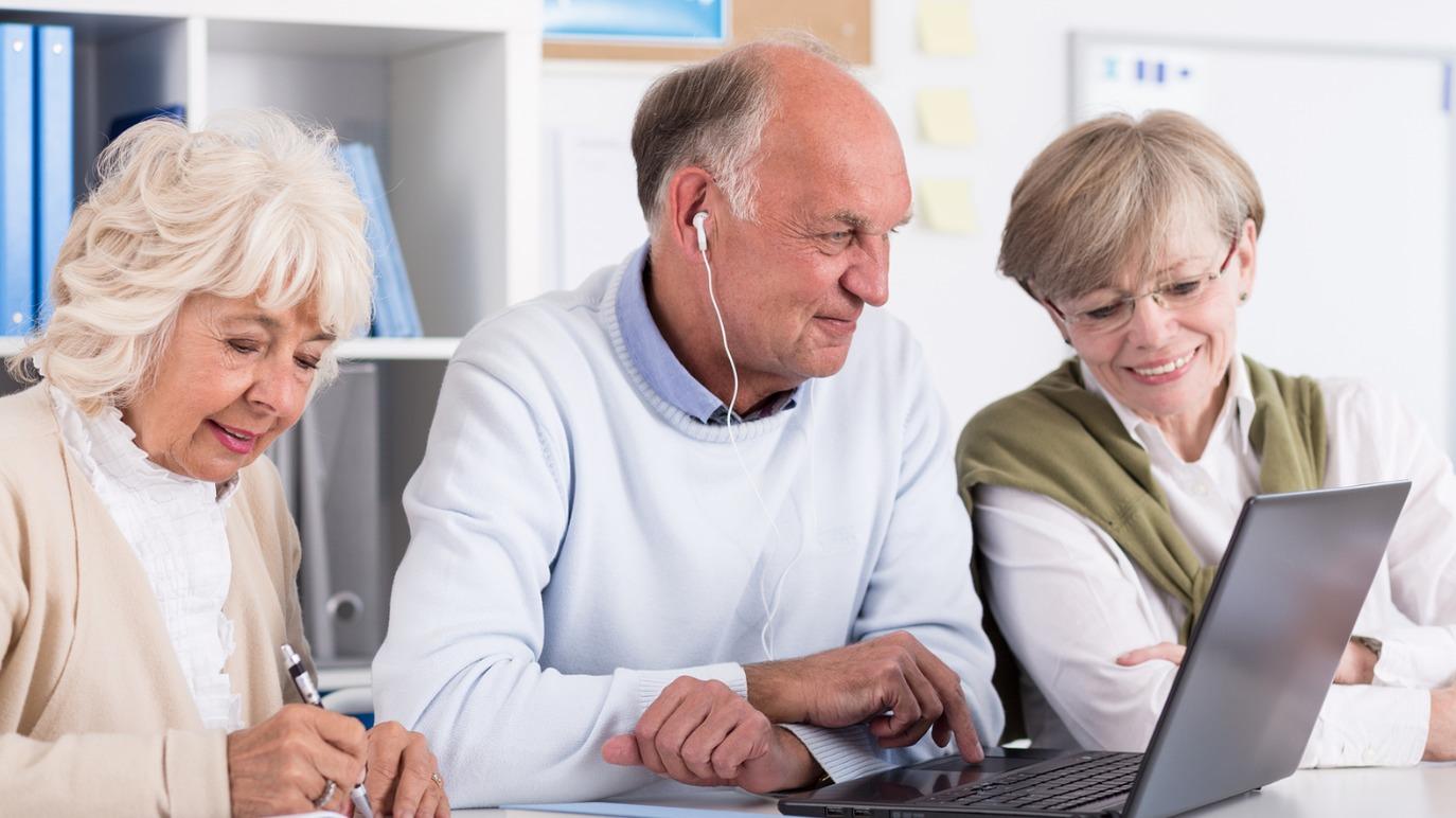 Tre ældre mennesker sidder sammen og ser på en bærbar computer, mens de smiler og lytter til musik.
