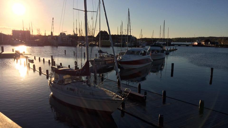Solnedgang over en havn med sejlbåde og motorbåde fortøjet ved pæle i vandet.