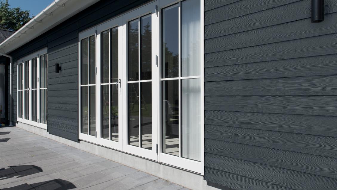 Moderne husfacade med grå træbeklædning og store hvidrammede vinduer.