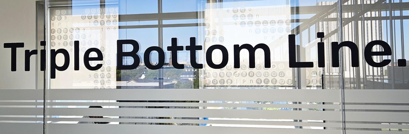 Skilt med teksten "Triple Bottom Line" på en glasvæg i et kontormiljø.
