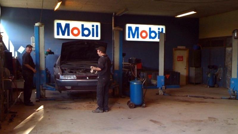 To mekanikere arbejder på en bil i en værksted med Mobil-skilt. Der er værktøj og udstyr synligt.