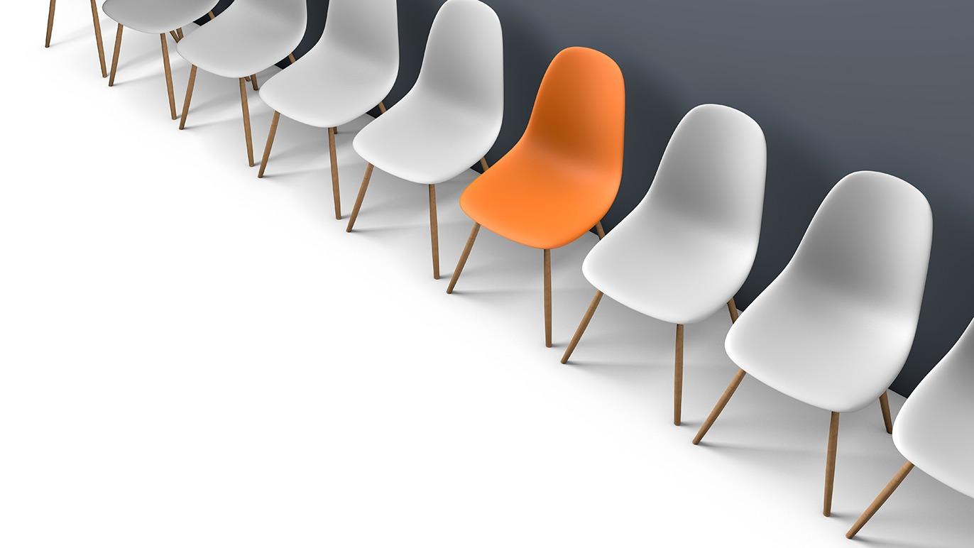 En række moderne stole i hvid og en orange stol, der skiller sig ud mod en grå væg.