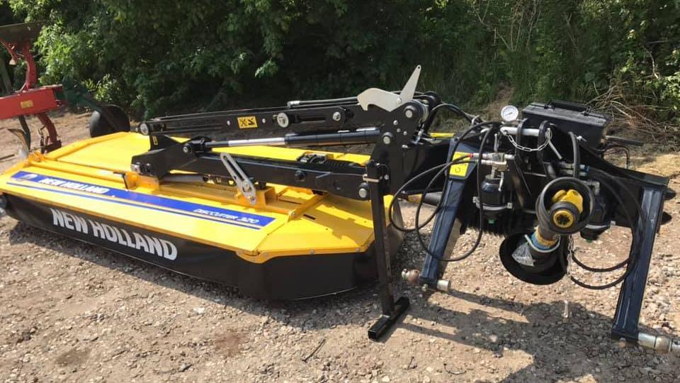 En New Holland græsslåmaskine med hydrauliske funktioner, placeret på en grusvej omgivet af grøn vegetation.