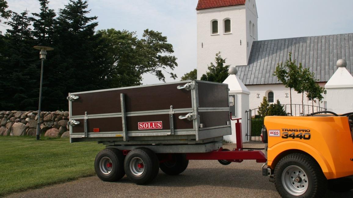 En trailer parkeret foran en kirke med en grå bygning og grønne områder omkring.