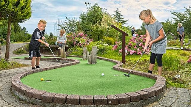 Børn spiller minigolf på en grøn bane omgivet af blomster og træer en solrig dag.