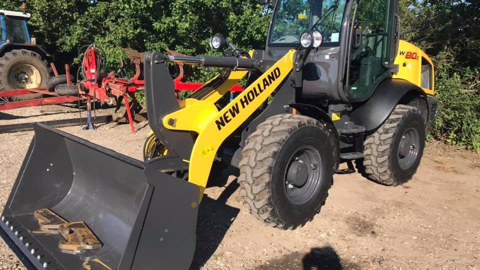 En New Holland minilæsser med skovl står på en grusvej omgivet af grønne træer.