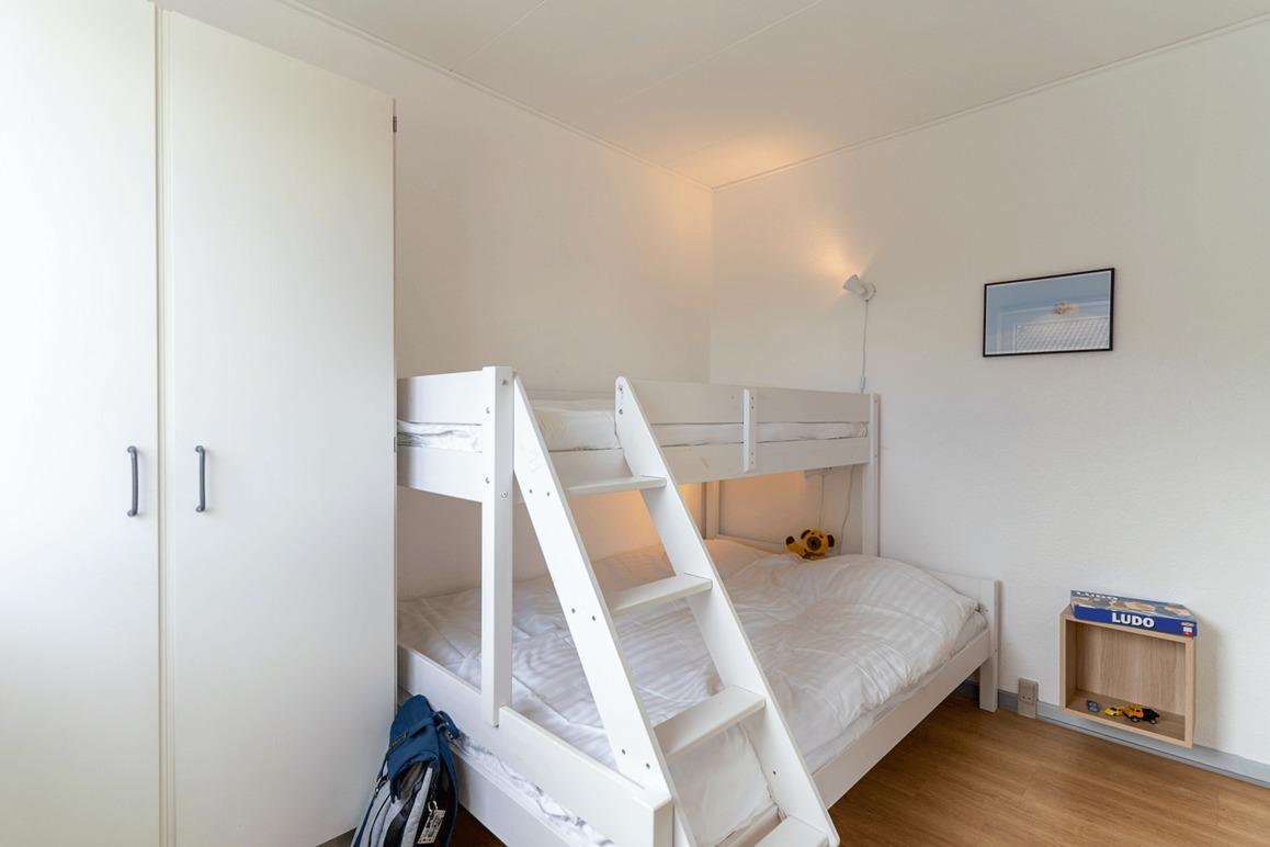 Lille værelse med en køjeseng, hvidt sengetøj og en kommode ved siden af. Der er også en lampe og et billede på væggen.