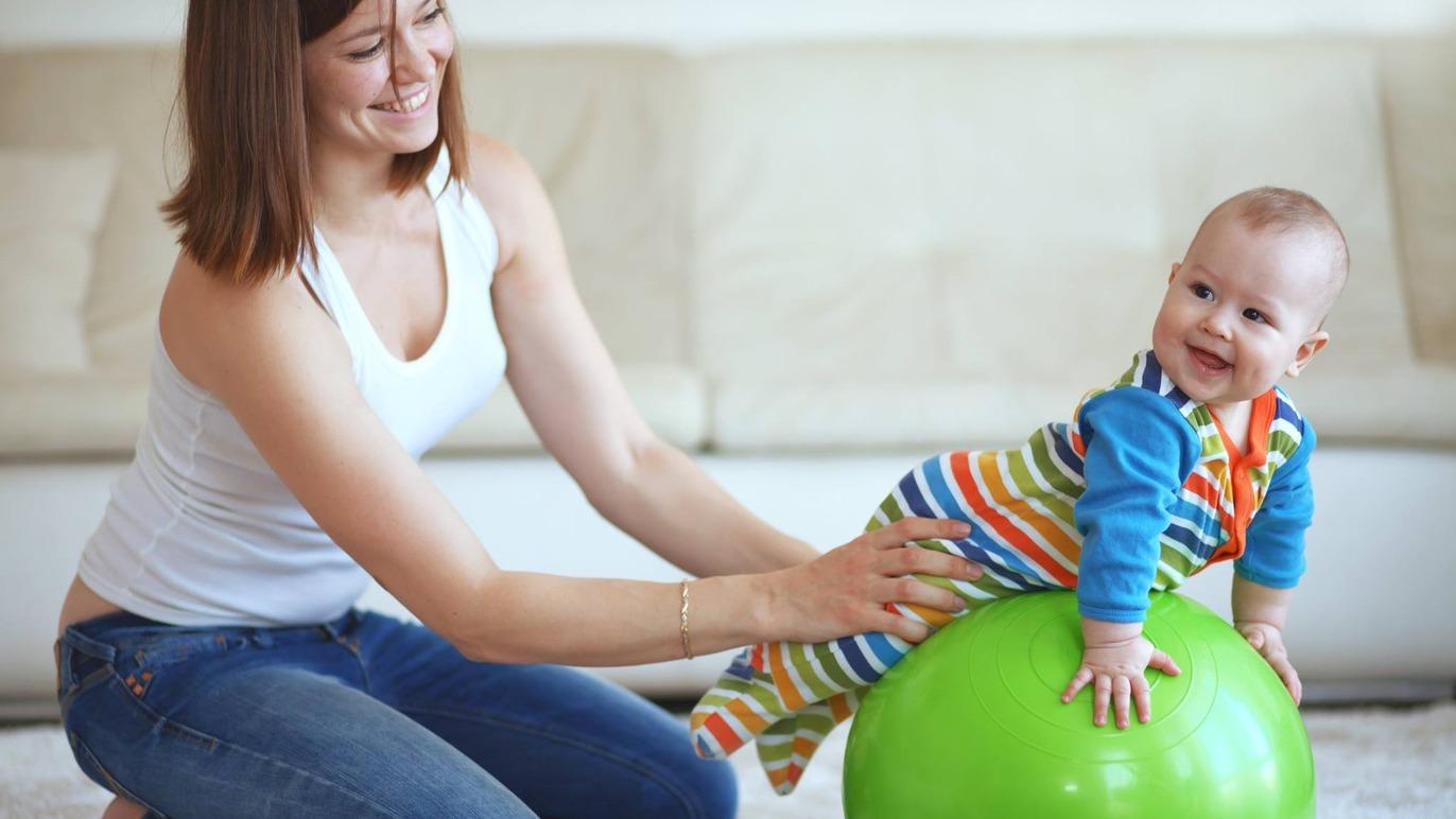 Mor leger med sin baby på en grøn bold i stuen, mens de begge smiler og har det sjovt.