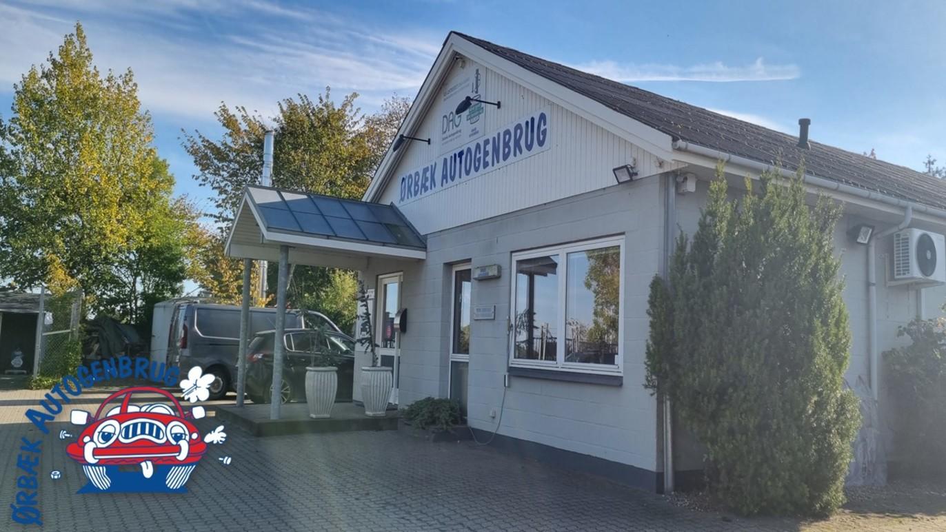 En bygning med solpaneler og en parkeret bil foran, omgivet af træer og en blå himmel.