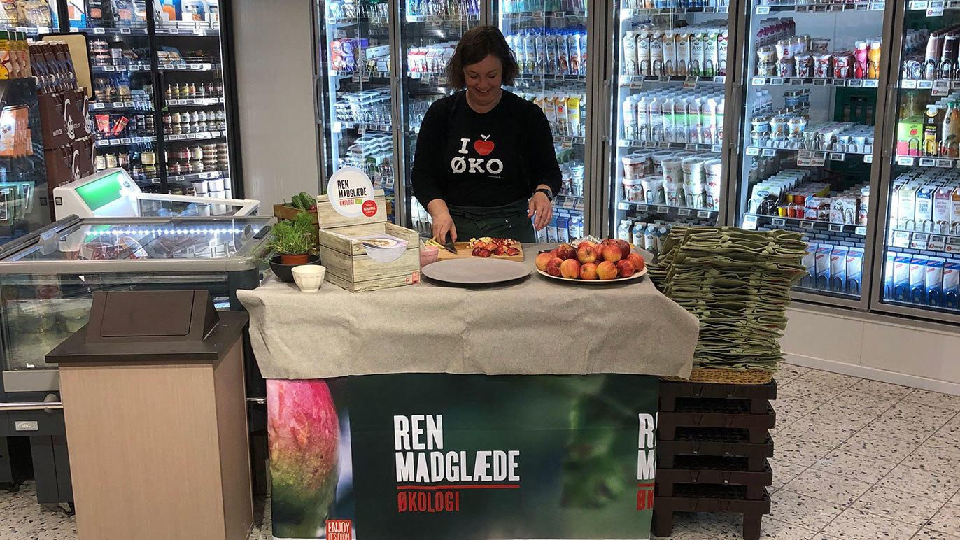 En kvinde står ved en bod med frugt og grøntsager i en butik, omgivet af køleskabe med drikkevarer.