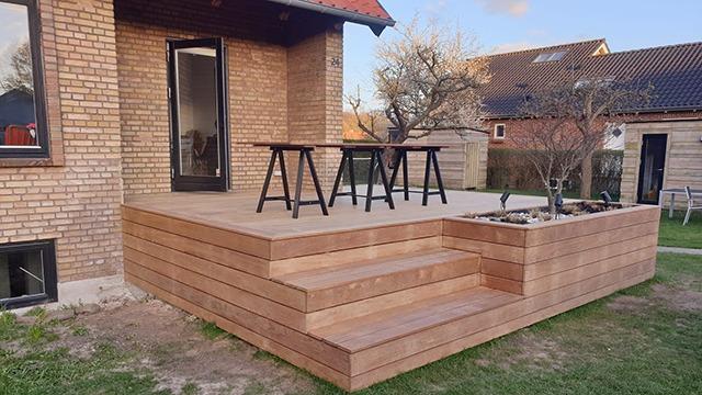 Terrasse i træ med trapper og borde foran et hus, omgivet af græs og træer.