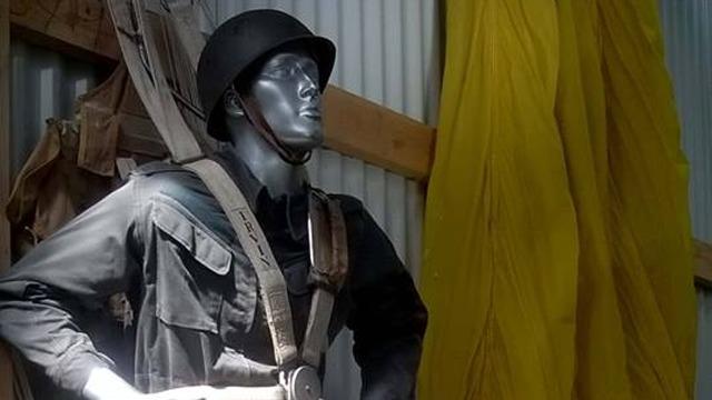 Mannequin iført militæruniform og hjelm, stående i et rum med gule gardiner.