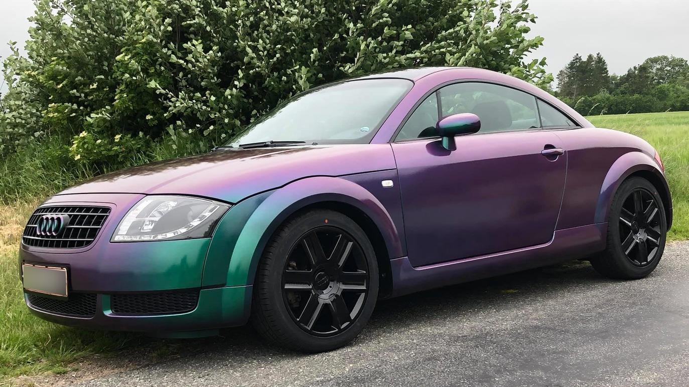 En farverig Audi TT med en gradient af lilla og grøn farve, parkeret ved en græsmark.