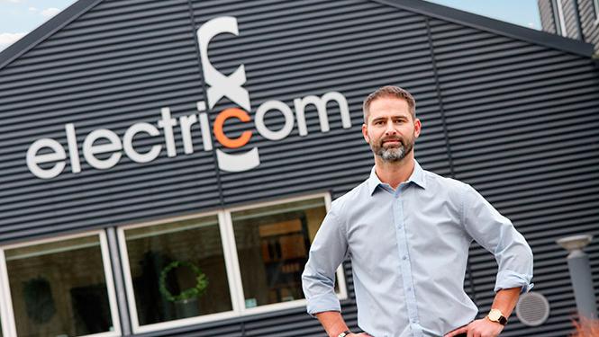 Mand står foran en bygning med logoet for electricom, iført skjorte og med hænderne på hoften.