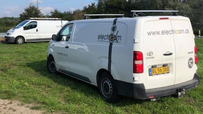 To biler parkeret på en græsmark, en hvid varevogn med logoet "electrom.com" synlig på siden.
