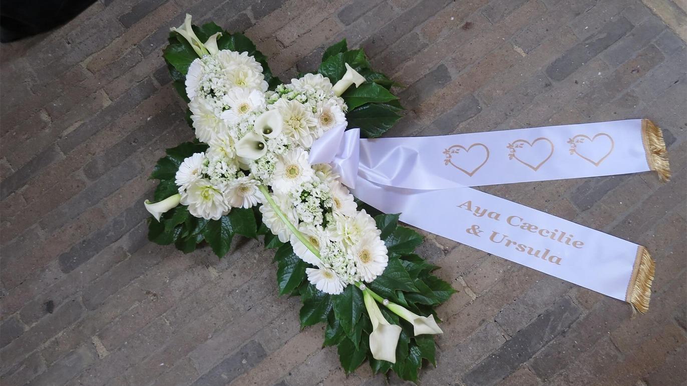 En hvid blomsterkrans formet som et kors med grønne blade og et bånd med navne.