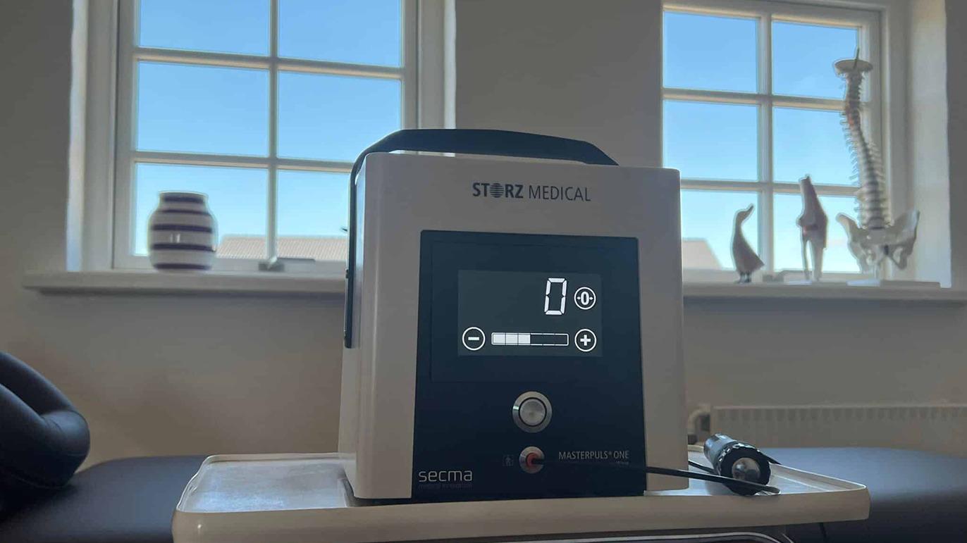 Medicinapparat med digital skærm og knapper, placeret ved vindue med blå himmel i baggrunden.