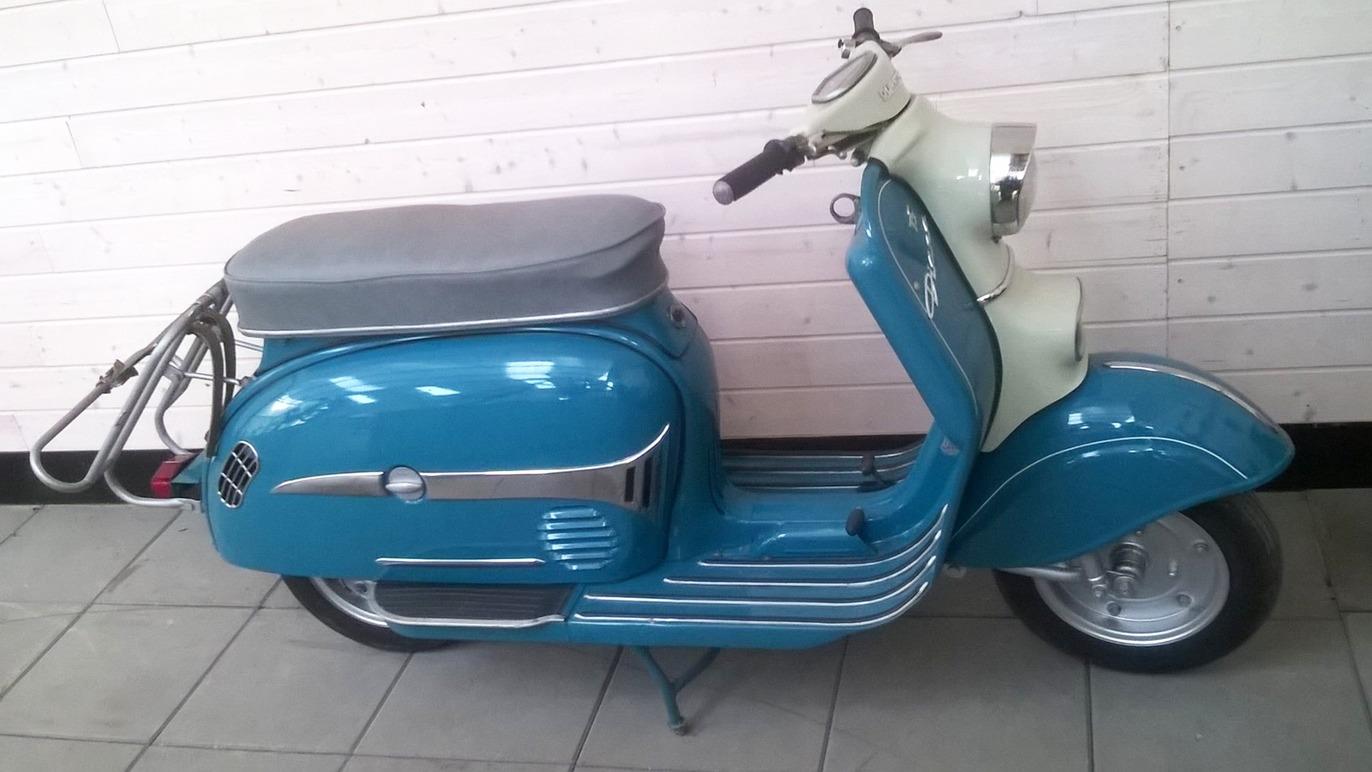 En vintage blå scooter med grå sæde, parkeret på fliser i et lyst rum.
