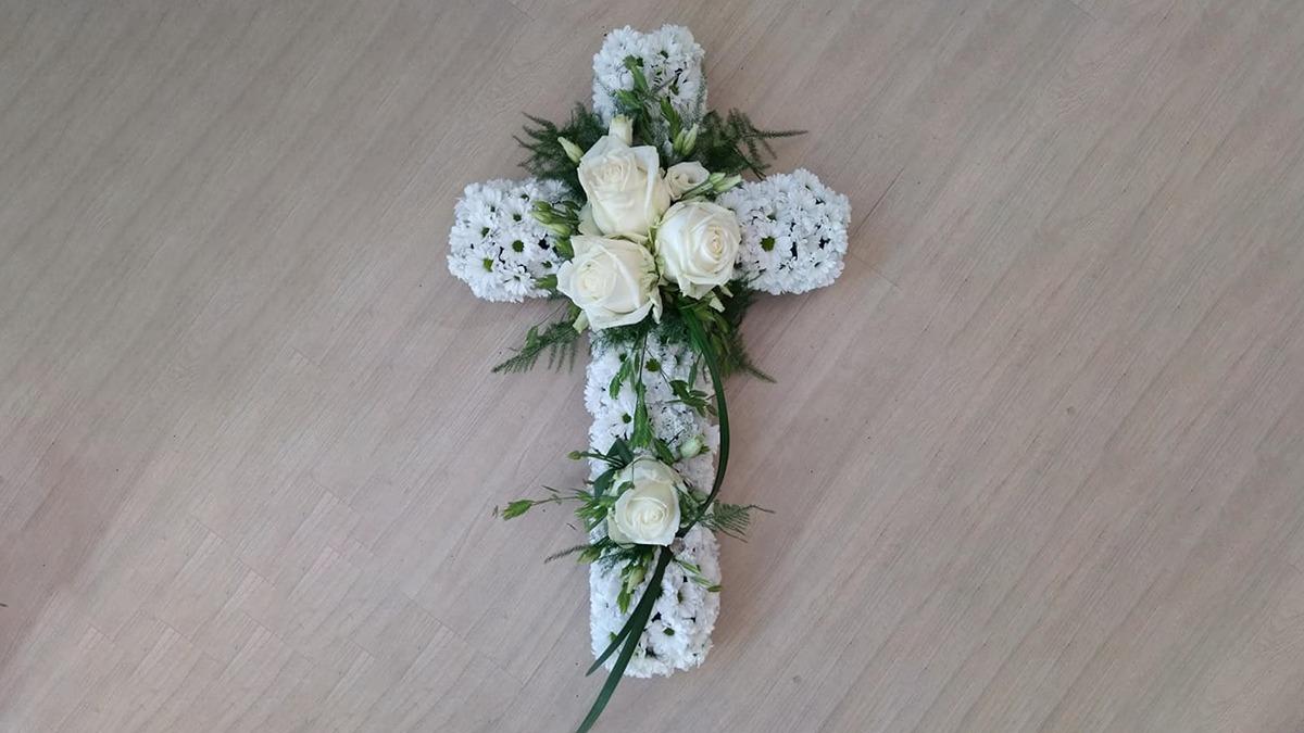 Hvid blomstercross dekoreret med roser og grønne blade på en lys træoverflade.