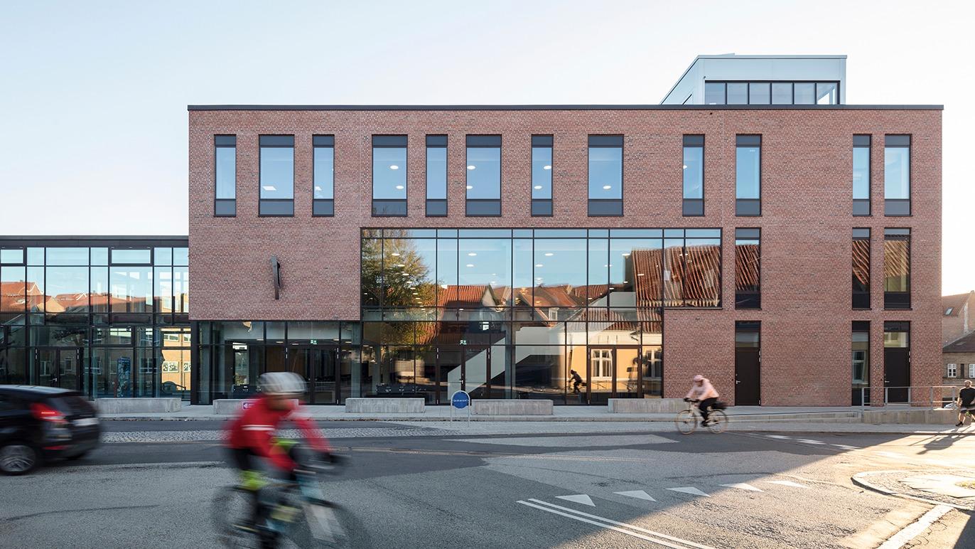 Moderne bygning med glasfacade og mursten, cyklister og biler på vejen foran.