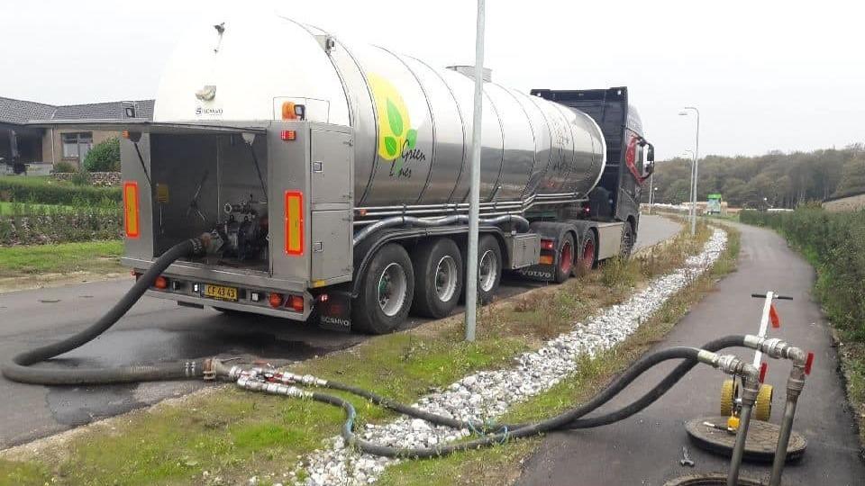 Lastbil med tank og slanger til affaldsopsamling på en vej ved en beplantning.