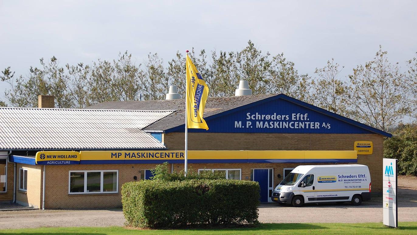 M.P. Maskincenter med gule og blå farver, bygning med flag og varevogn parkeret foran.
