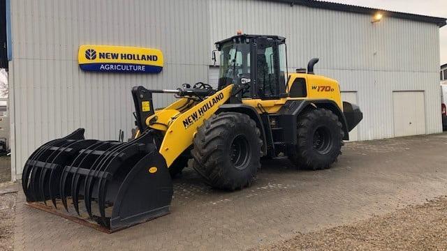 En gule New Holland landbrugsmaskine med skovl står foran en bygning med et skilt.