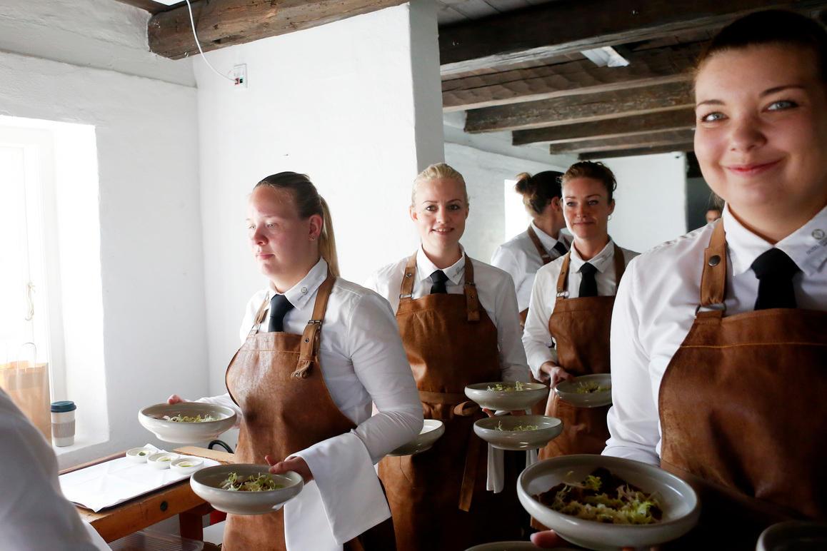 Fire tjenere iført brune forklæder bærer tallerkener med mad i en lys restaurant.