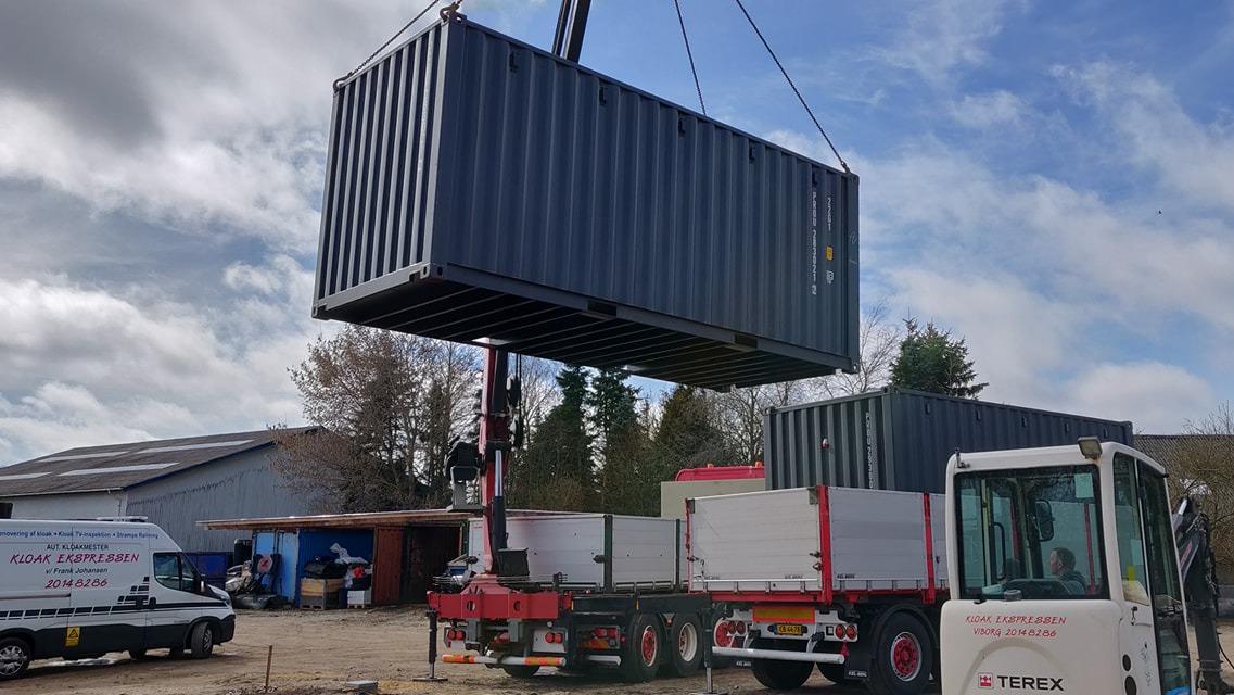 En kran løfter en stor container over en lastbil på en byggeplads med bygninger i baggrunden.