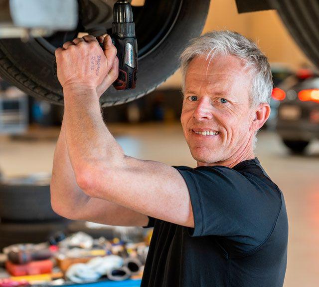 Mand arbejder med en elektrisk skruenøgle under en bil, mens han skifter dæk i et værksted.