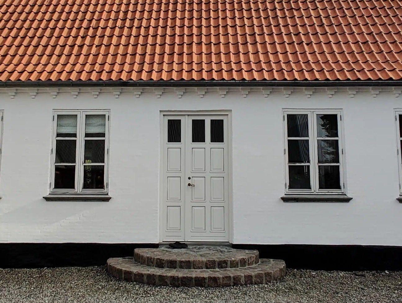 Hvidt hus med rød teglstens-tag og en dør med trin foran, omgivet af grus.