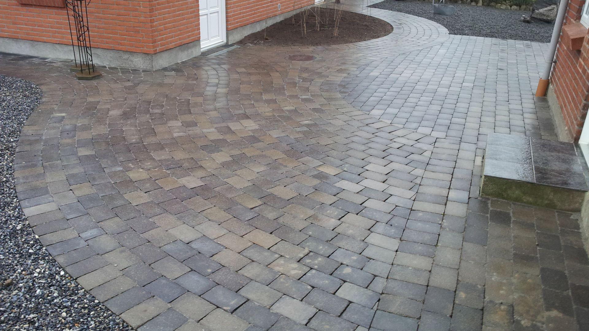 En nyanlagt indkørsel med brosten, der skinner efter regn, omgivet af grus og en murstensvæg.