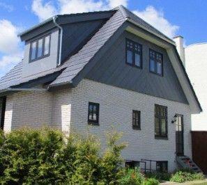 Et moderne hus med grå tag og hvid murstensvæg, omgivet af grønne buske og en blå himmel.