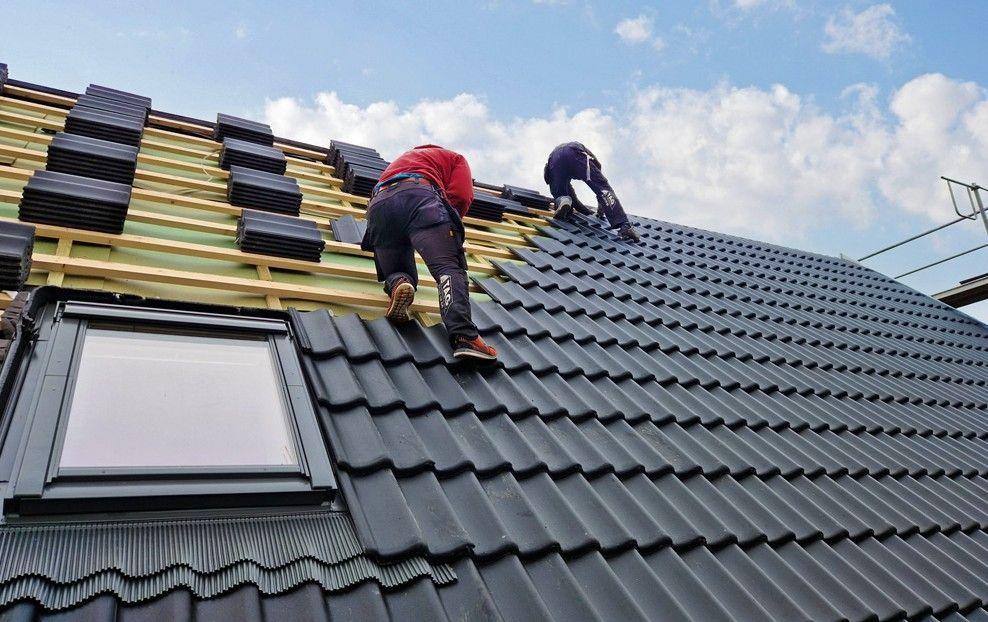 Tagdækkere arbejder på et sort tag med tagsten under en blå himmel med skyer.