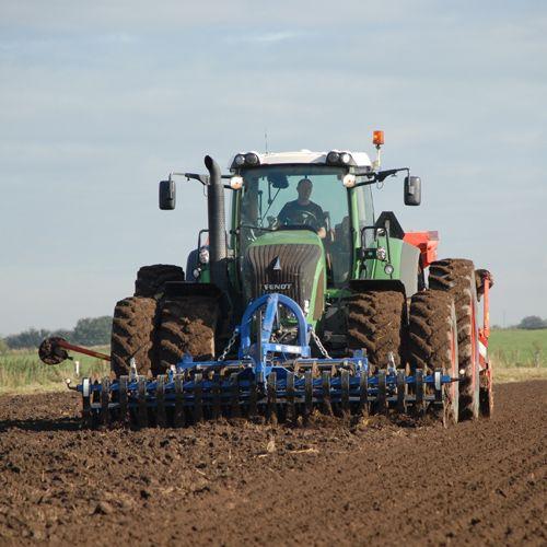 En traktor pløjer jorden på en mark med en blå plov under en klar himmel.