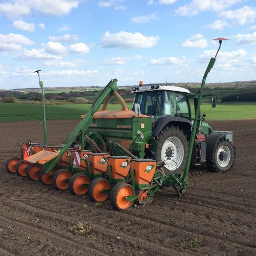 En traktor med en såmaskine arbejder på en mark under en blå himmel med skyer.
