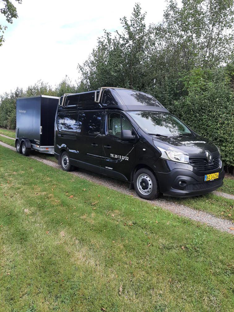En sort varevogn med trailer parkeret på en grusvej omgivet af grønne træer.