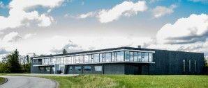 Moderne bygning med store vinduer og græsplæne, beliggende ved en vej under en blå himmel med skyer.