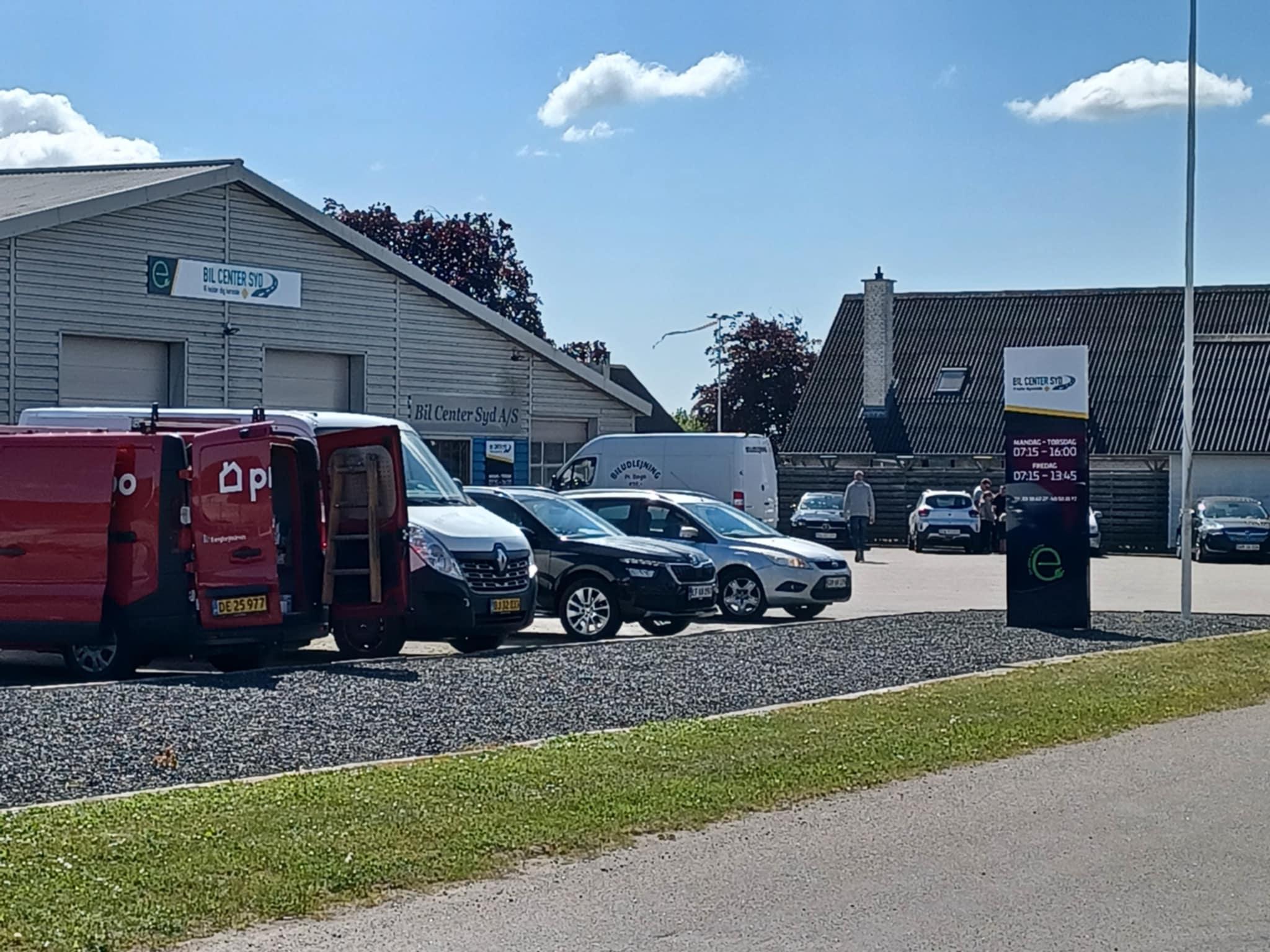 Billede af en virksomhed med flere biler og varevogne parkeret foran bygningen på en solrig dag.