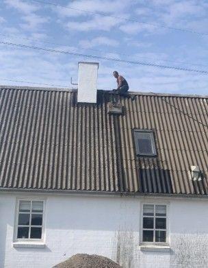 Mand arbejder på taget af et hus med skrå tag og en skorsten, mens der er blå himmel i baggrunden.