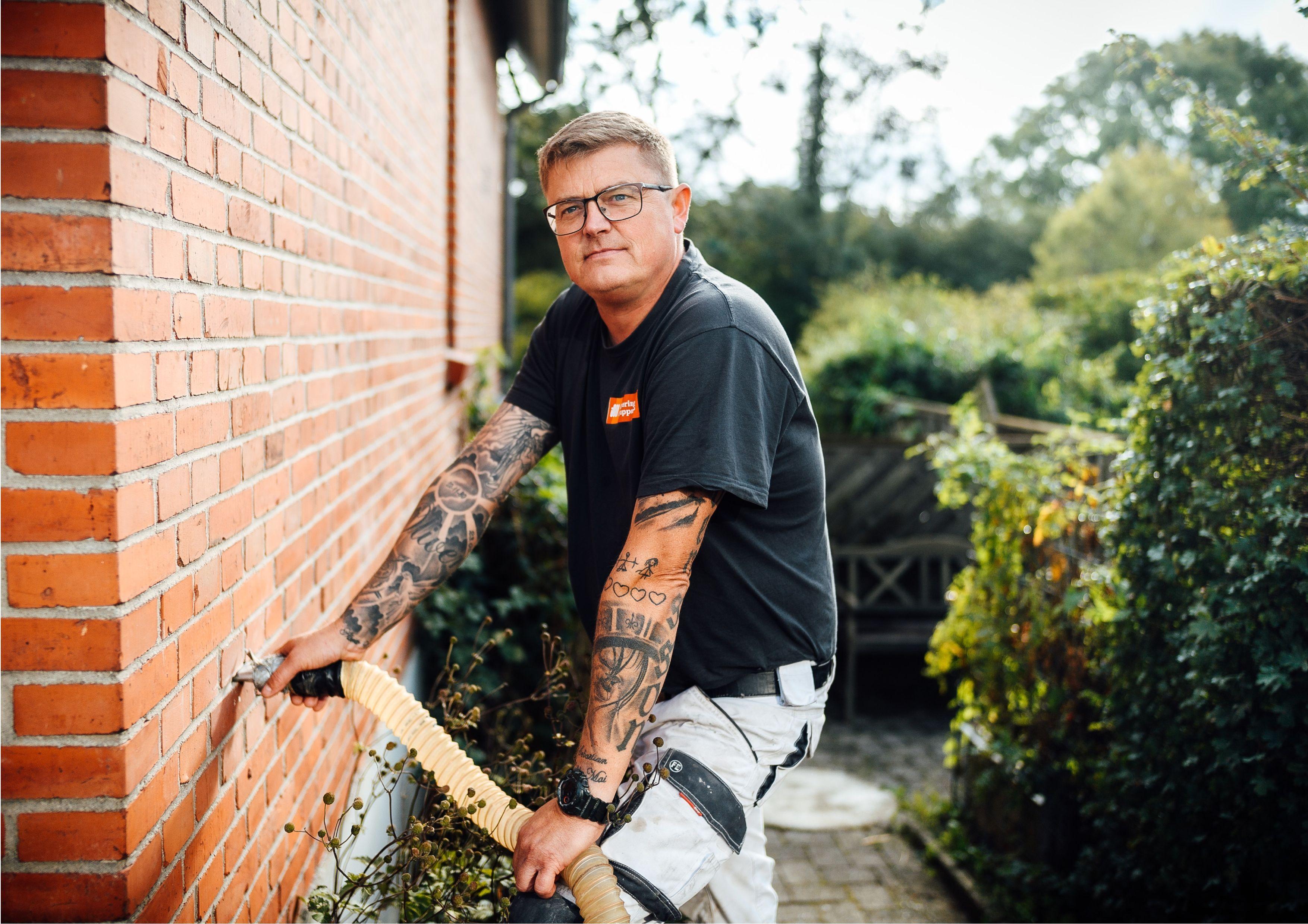 Mand med tatoveringer arbejder på murværk i haven. Solrigt vejr og grønne planter i baggrunden.