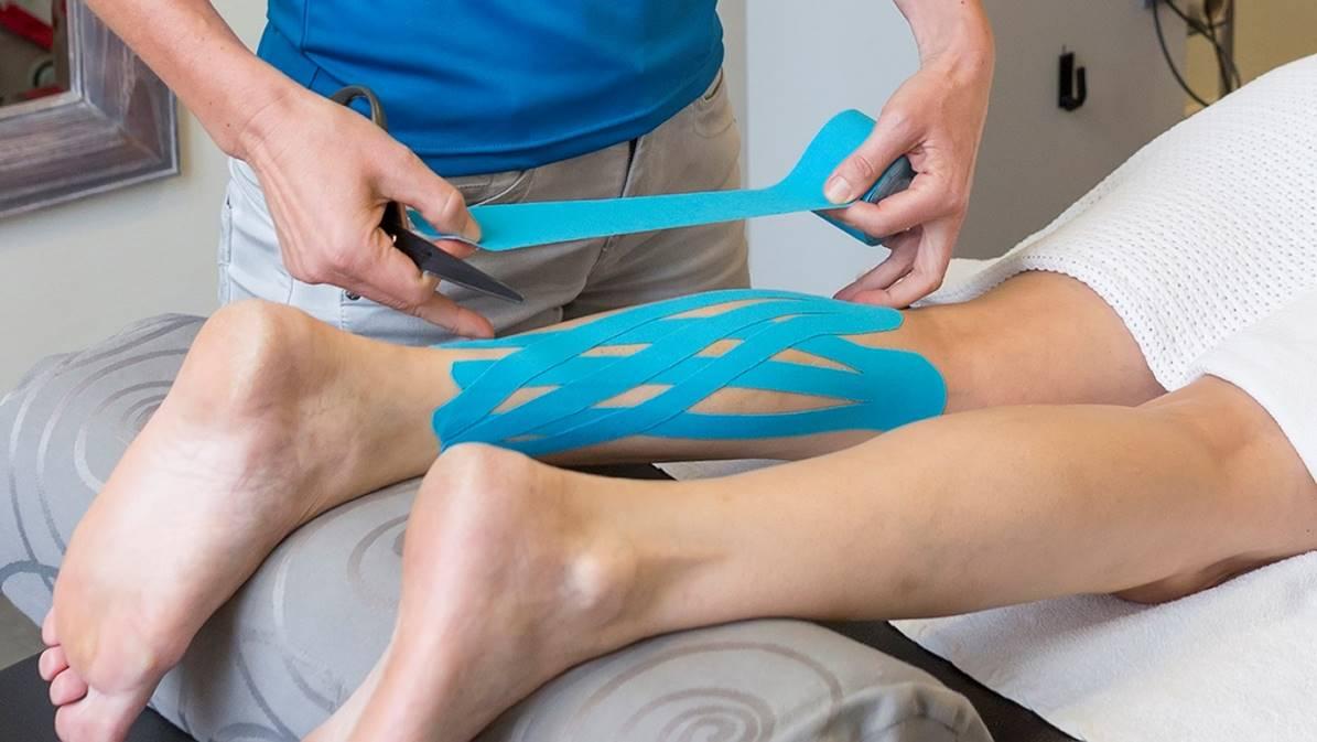En terapeut påfører blå kinesiotape på en patients ben under en behandling.
