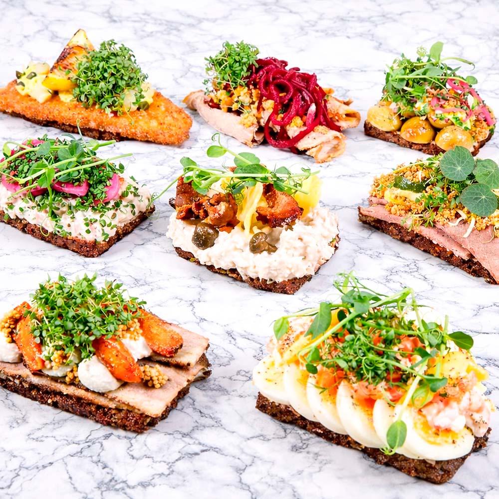 Et udvalg af smørrebrød med forskellige toppings som fisk, kød, grøntsager og urter på rugbrød.