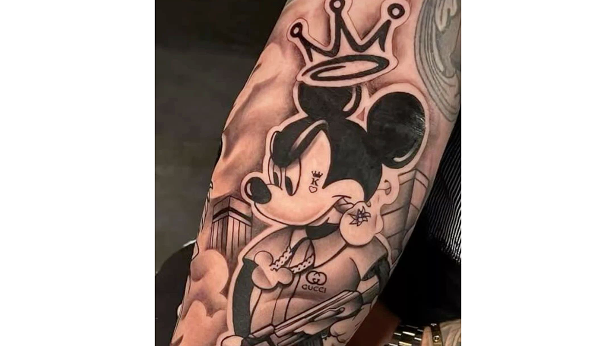 Mickey Mouse tatovering med krone og guitar på en arm, detaljeret sort-hvidt design.