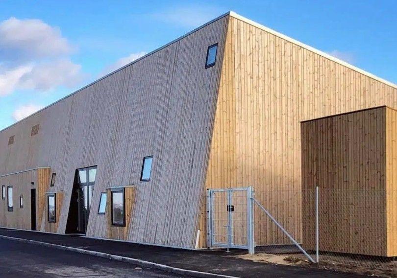 Moderne bygning med træfacade og skrå vægge, beliggende ved en vej under en blå himmel.